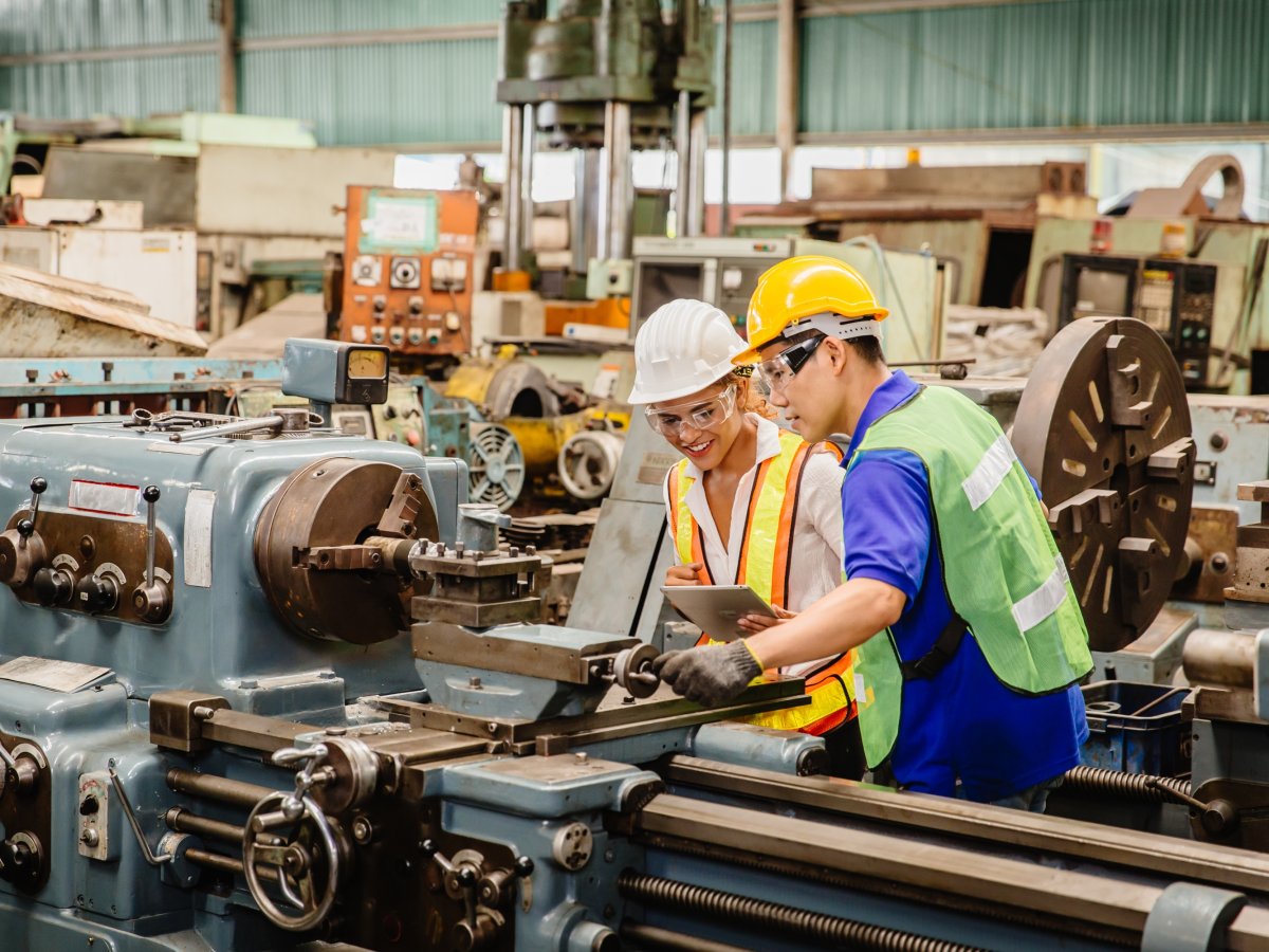 Two workers wearing helmets inspecting a machine in a factory hall.